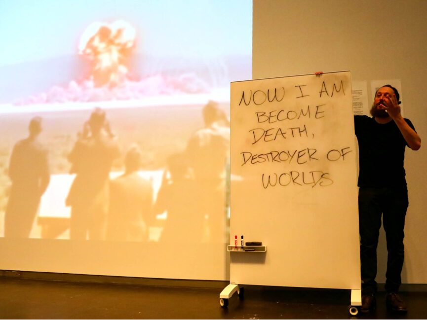 A projection of a nuclear explosion is in the background of a performer at a whiteboard that reads "Now am I become death destroyer of worlds"