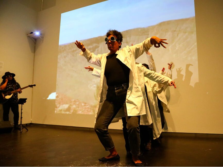 Performers wearing lab coats perform in front of a projection of a desert landscape.