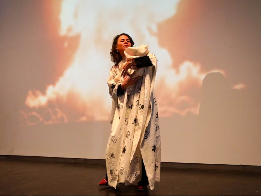 A performer appearing to hold a baby performs in front of a projection of a nuclear explosion