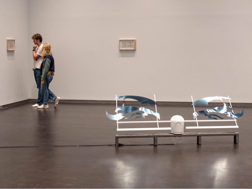 A mechanical sculpture sits on the floor of a gallery as visitors look artwork on the walls