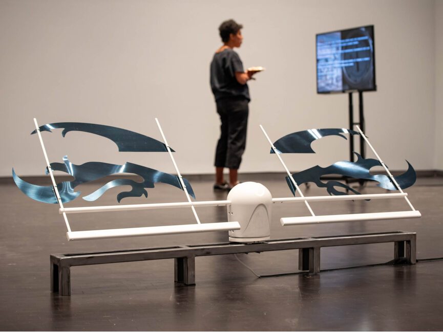 A mechanical sculpture sits on the floor of a gallery as a visitor watches a video monitor in the background