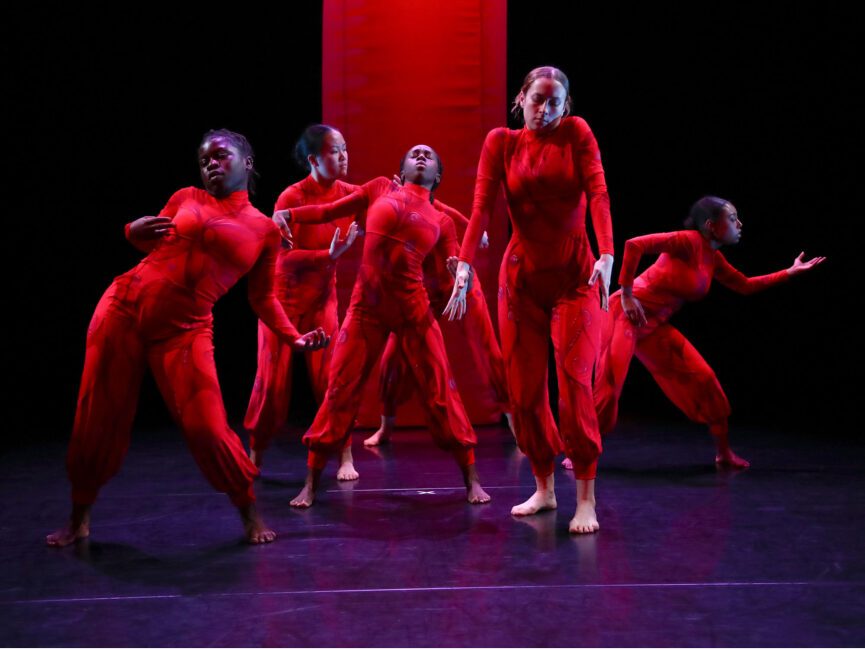 Six dancers on a dimly lit stage, all dressed in dark red, strike various poses in a tight grouping.