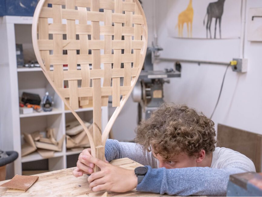 A student artist tries to balance the base of sculpture that appears to be a tennis racket on workbench.