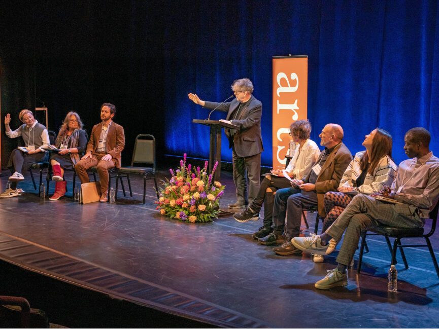 Poets sit in chairs on a stage as Paul Muldoon stands in the center to introduce the event