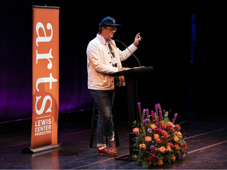 A person stands at a podium in front of a banner that reads arts and behind a boquet of flowers.