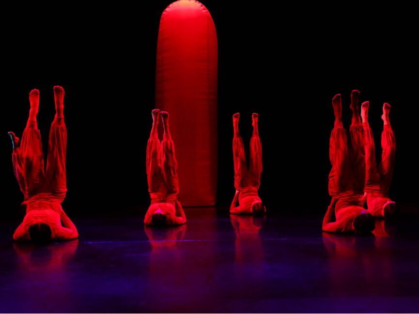 A line of 5 dancers lie on the floor with their legs kicked upwards