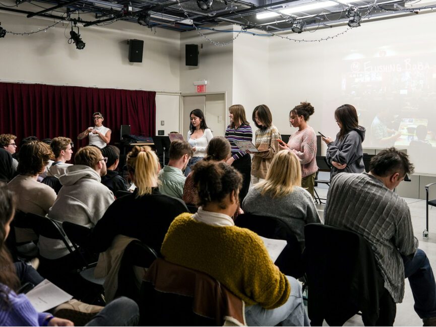 An audience watches students standing in front of a room reading from scripts