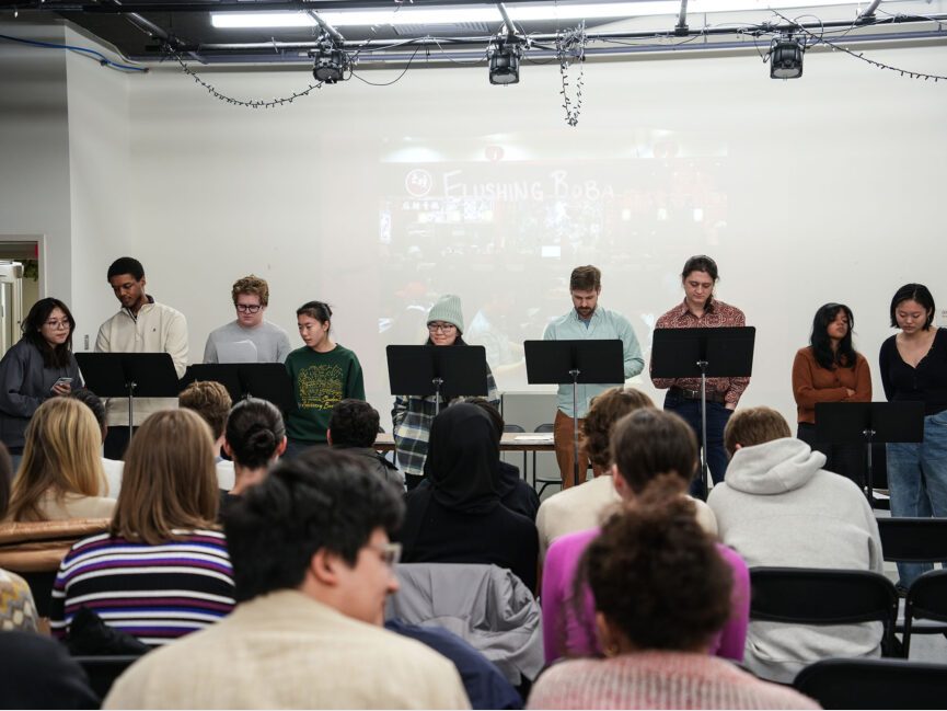 An audience watches students standing in front of a room reading from scripts