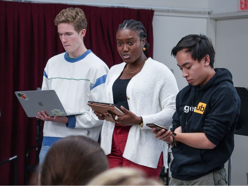 Three people stand and read from laptops and phones