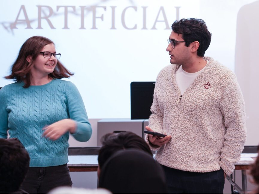 Two people read from their phones and perform in front of a projection that reads "Artificial"