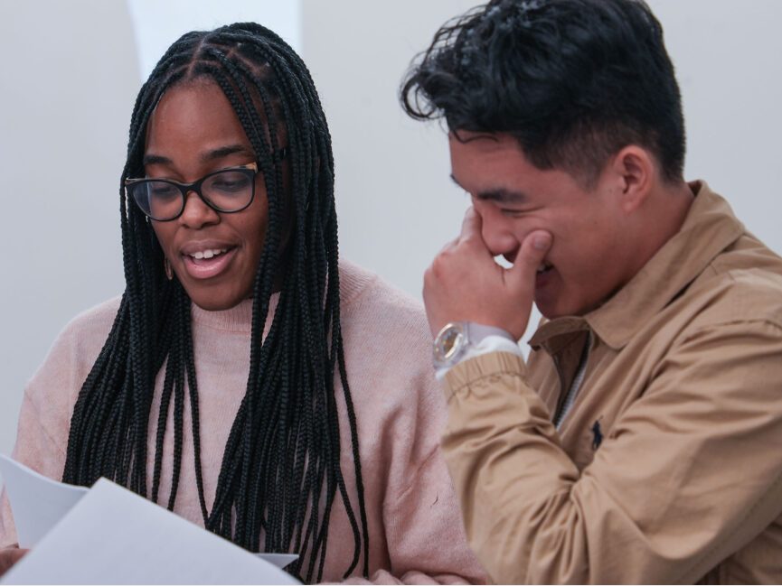 Two people read from a script. One of the readers tries to hold bacl laughter