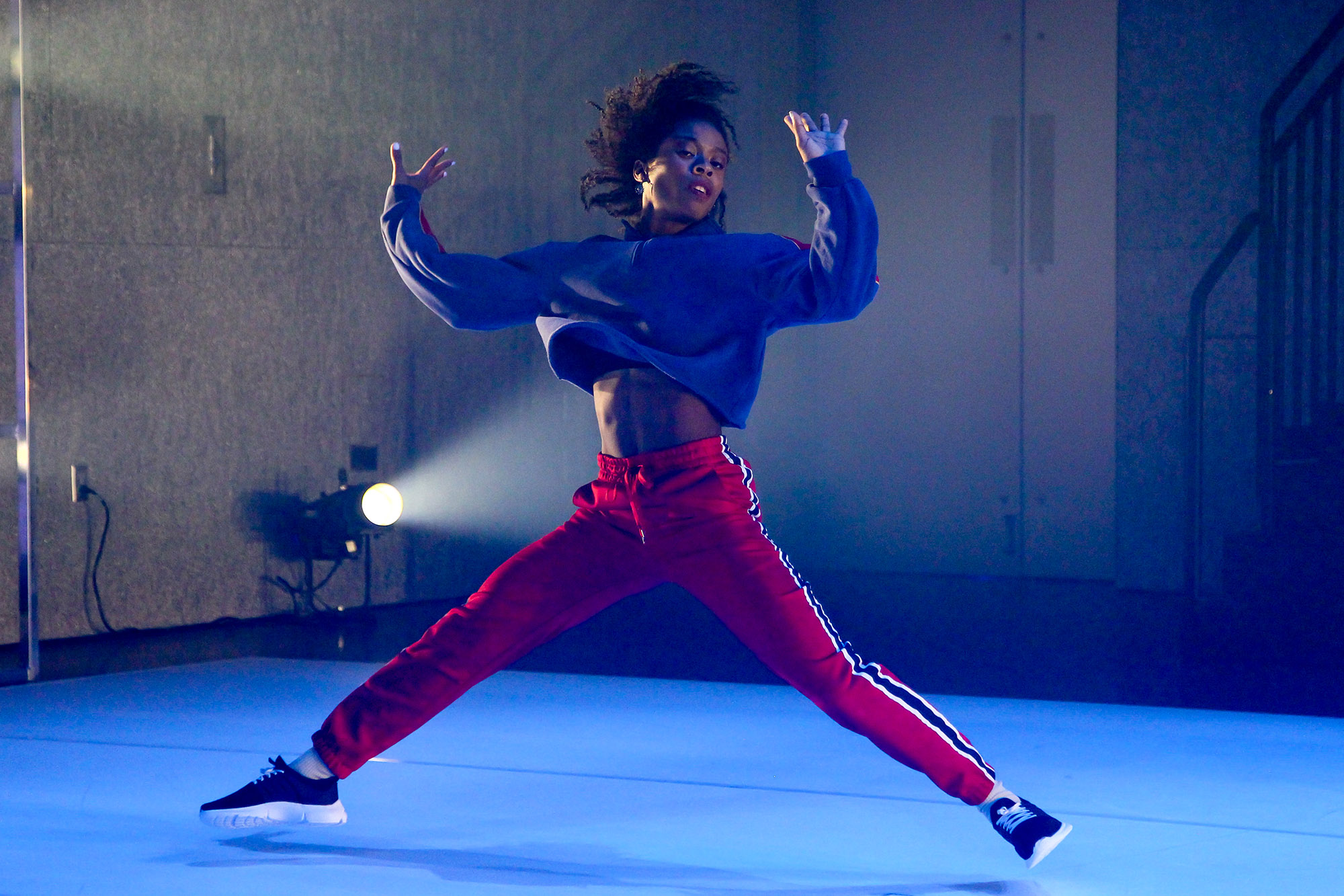 Runako Campbell lunges in mid-air with legs and arms spread wide. She wears red sweats and a navy blue hoodie.
