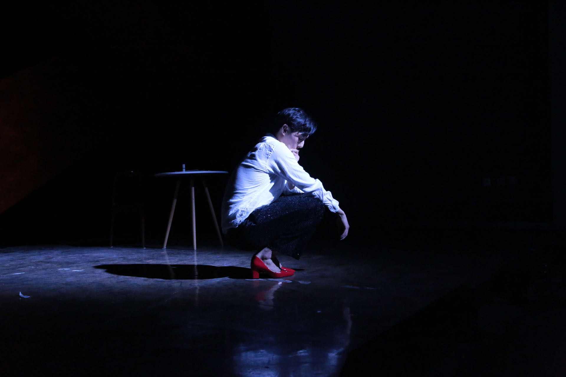 An actor squats on a dark stage in spotlight, wearing red heels, dark pants and a whie shirt.