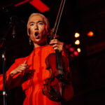 Yuniya edi kwon sings with a bold expression and holds a violin while wearing a bright red dress.