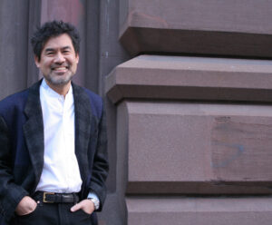 David Henry Hwang smiles and stands near a stone column, with his hands in his pockets.