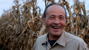 A person stands smiling in front of the tall stalks of a cornfield.