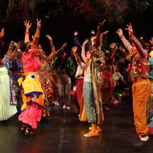 A large group of performers in colorful costumes dance and sing with enthusiasm on stage