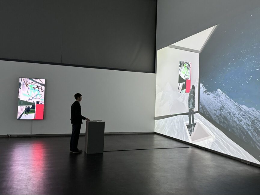 A person stands at a podium in front of a large projection in a dark gallery