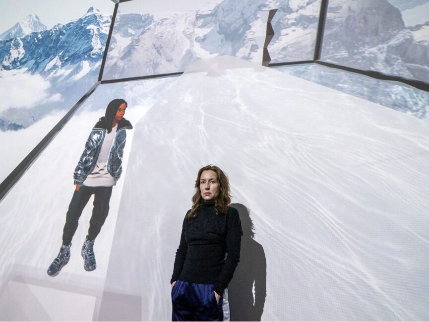Artist Josephine Meckseper poses in front of a digital projection of a figure standing in a virtual space