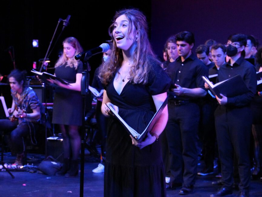 A singer holding a song book performs in front of a chorus on stage