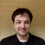 David Boyd smiles while looking off to the right, standing near a yellow brick wall.