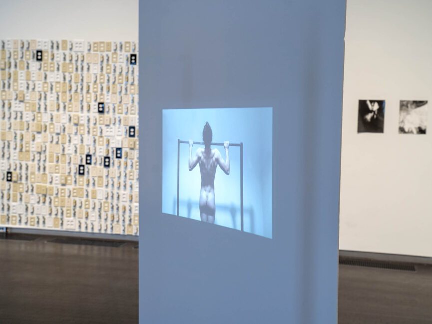 A video of a nude person doing pull ups is projected on a screen in an art gallery