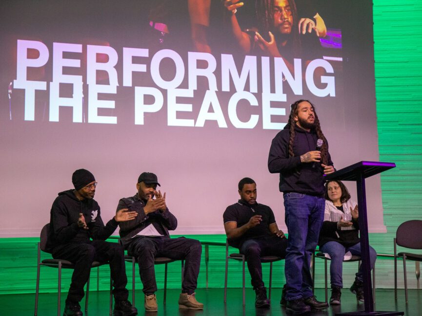 A person holding a microphone addresses an unseen crowd as other sit in chairs in front of a projection screen that says "Performing the Peace"