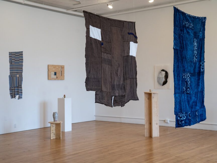 Two large pieces of fabric art hang from the ceiling in a gallery space with artwork on the walls