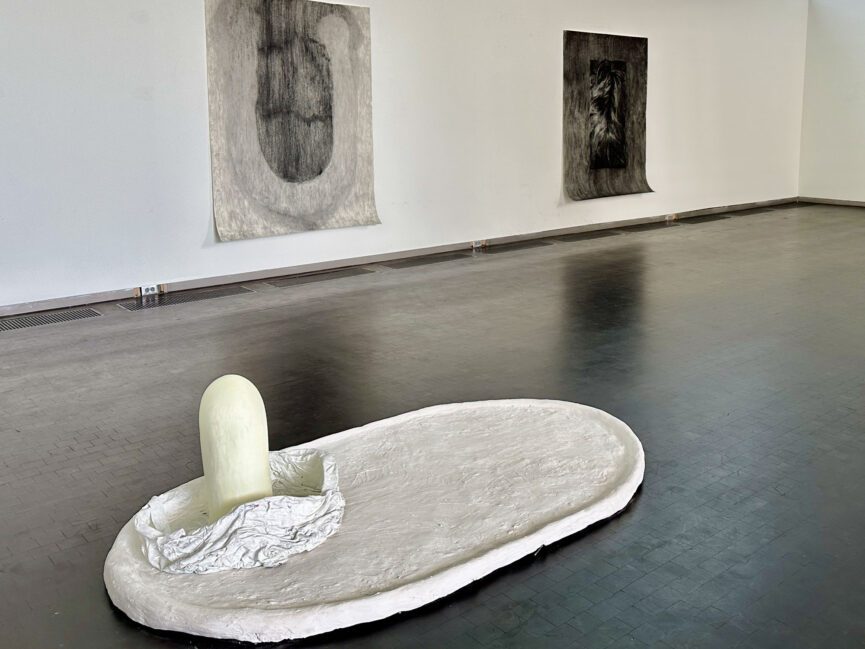Charcoal works on paper hang on a gallery wall behind a sculpture sits on the floor