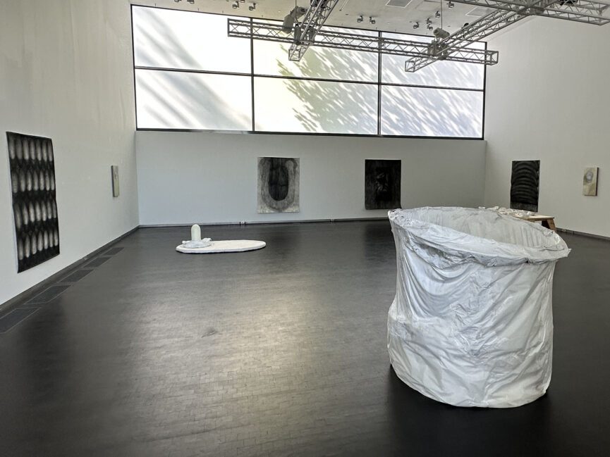 A large painted white cylindrical sculpture sits at center while a charcoal drawing hangs on wall behind.