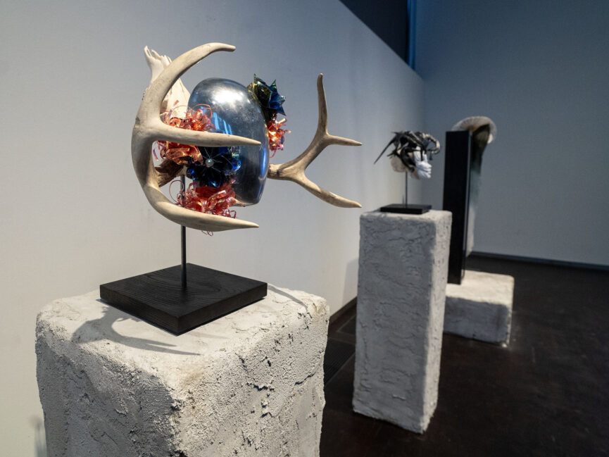 A reflective mask with antlers sits on a pedestal in a gallery