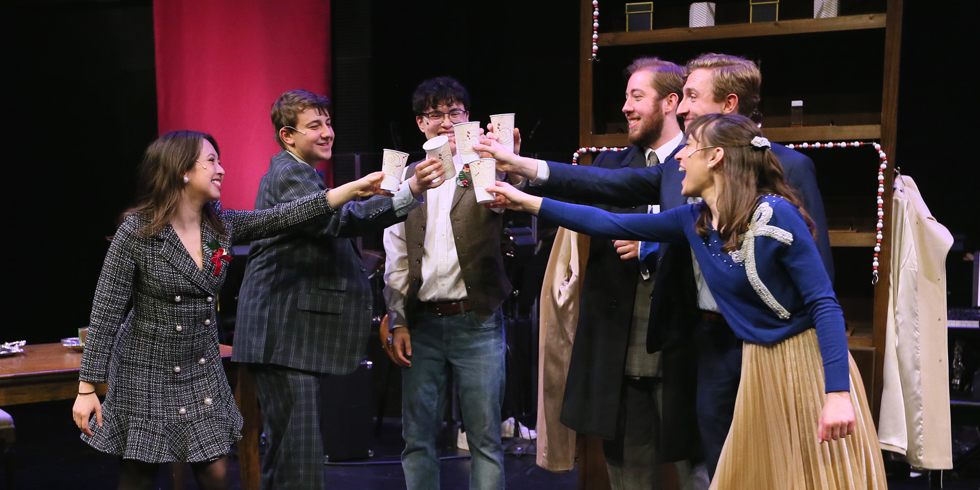 Six actors and actresses in a circle raising their glasses up to 'cheers' together