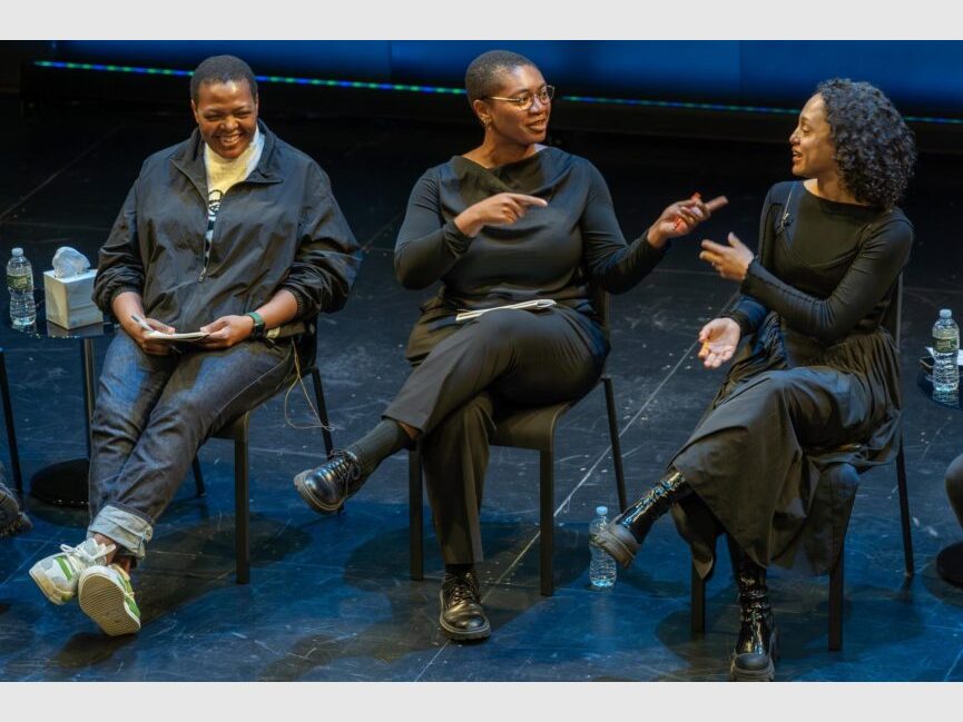 3 people talk and gesture while seated in chairs.