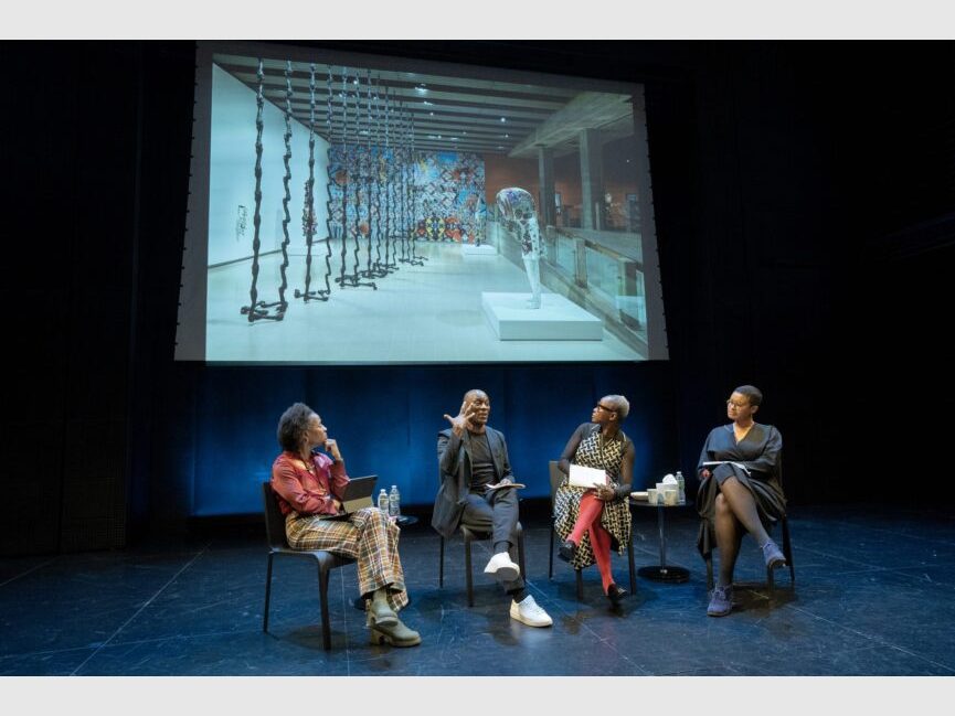 4 people seated in chairs talk and gesture to a large screen behind them.