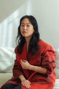Gi gazes at the camera and sits on a white couch wearing a red blazer.