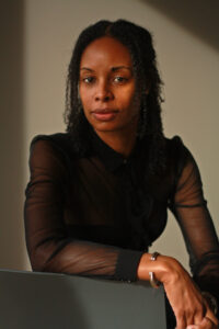 Nicole Sealey gazes at the camera, seated with arm resting on the back of a chair.