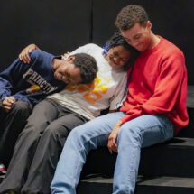 Three actors sit on a step and embrace