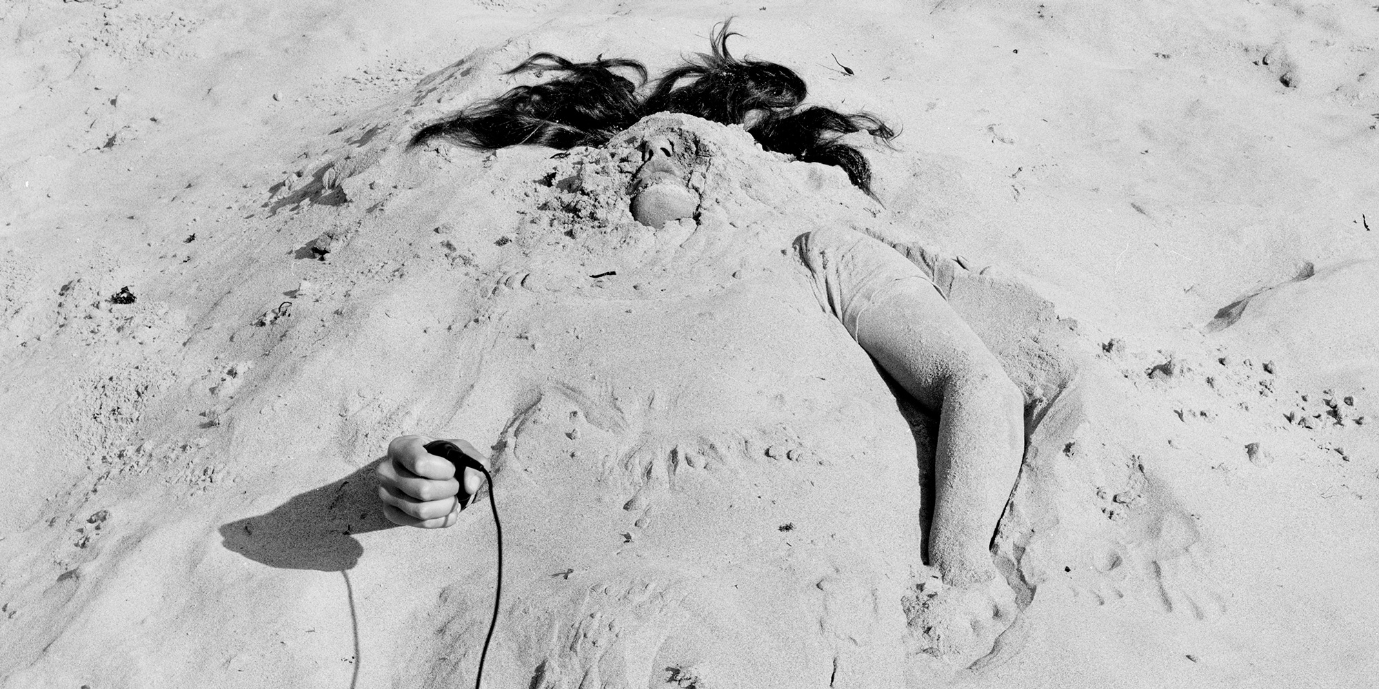 A person is lying buried in the sand with both arms, some hair, and nose and chin partially uncovered.