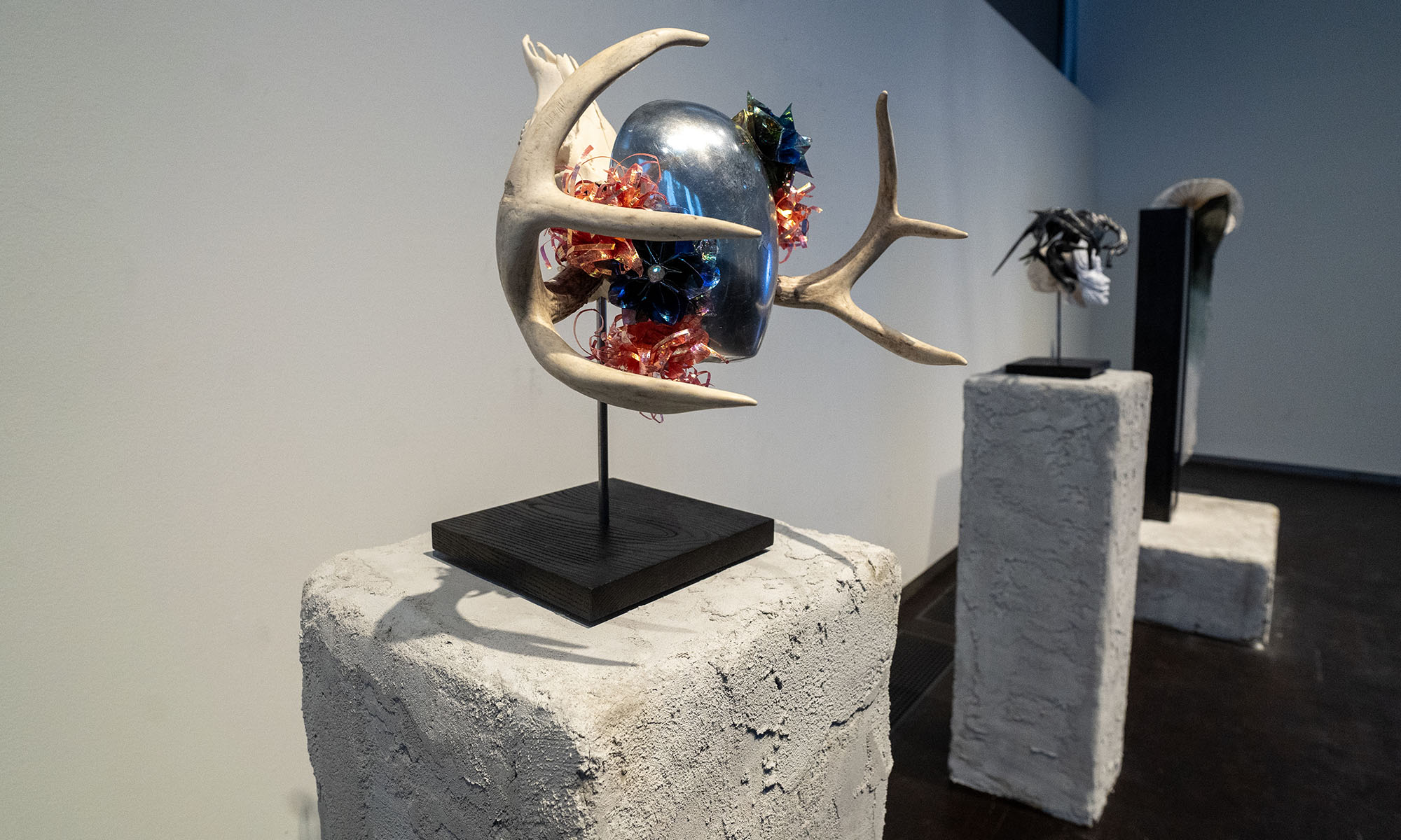 A reflective mask with antlers sits on a pedestal in a gallery