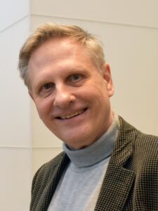Robert Spoo smiles, wearing a grey turtleneck shirt and brown checkered blazer.