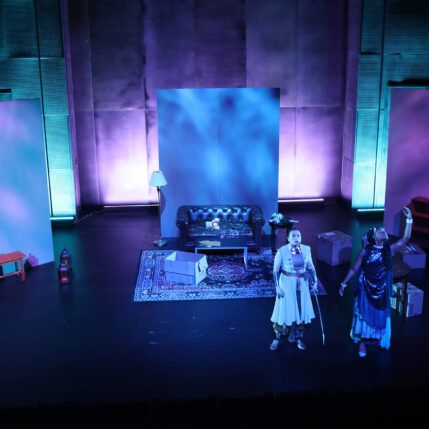 Stage set with a sofa, cardboard boxes, actors in costume, and blue and purple lighting.