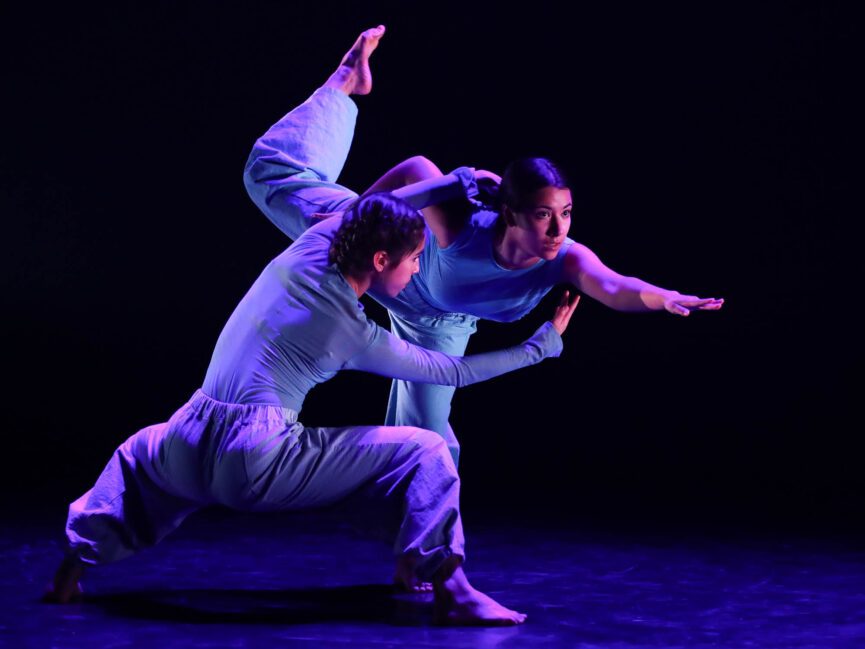 Two dancers in blue perform on a dimly lit stage, one crouching and the other balancing on one leg.