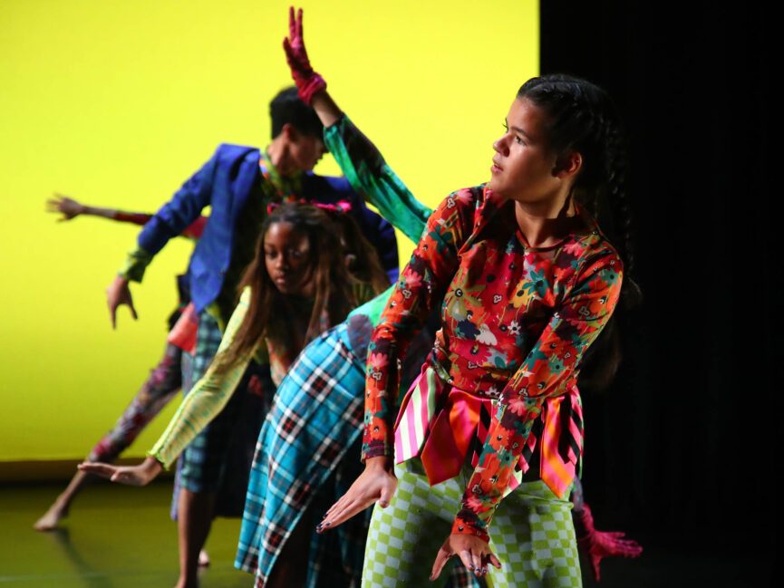 Dancers in colorful costumes performing on stage with a bright yellow backdrop.