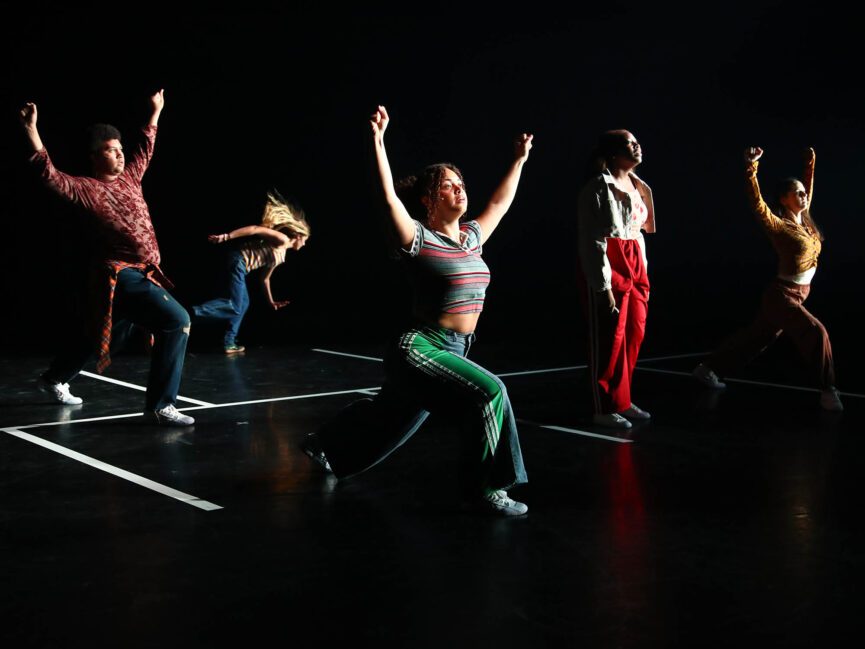 Five dancers perform dynamic poses on a dark stage with geometric white lines.