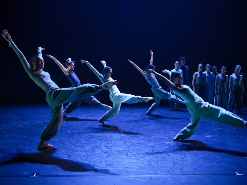Dancers perform dynamic poses under blue stage lighting, with a group in the background.