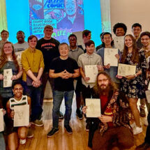 A group of people pose with drawings in a gallery with a comic book poster projected behind them.