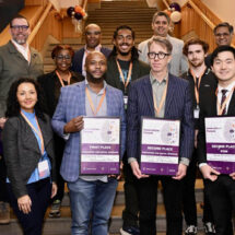 A group of people stand together on the bottom step of a staircase, some holding awards.