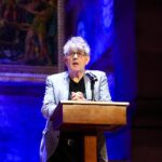 Paul Muldoon stands speaking behind a mic in a dark room lit with blue light.