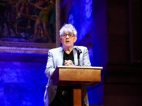 Paul Muldoon stands speaking behind a mic in a dark room lit with blue light.