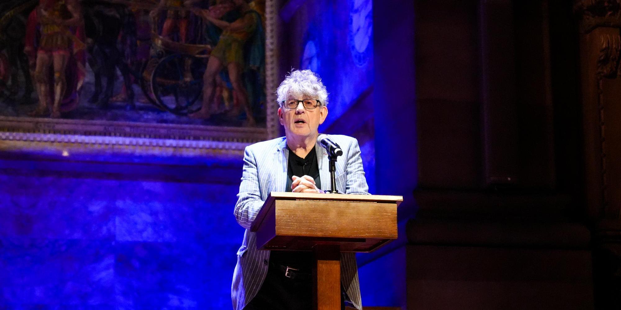 Paul Muldoon stands speaking behind a mic in a dark room lit with blue light.